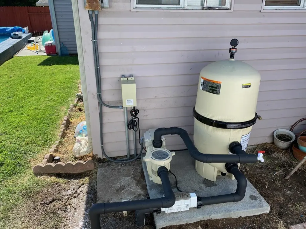 Cream filter tank and pump with black plumbing on a concrete pad beside the house