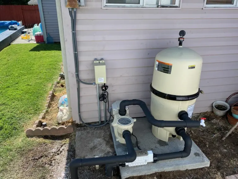 Cream filter tank and pump with black plumbing on a concrete pad beside the house