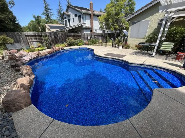 Freeform pool with a bright blue liner, boulders along one side and a pergola behind