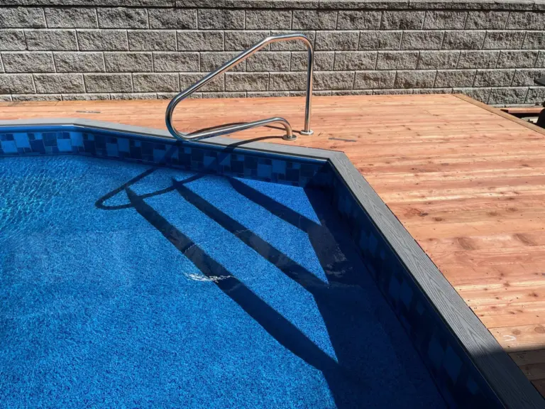 Pool with a blue mosaic tile border and a stainless ladder beside a redwood deck and a stone wall