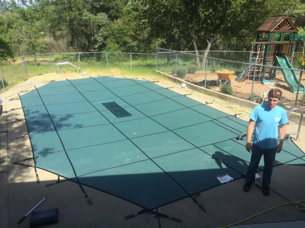 A green mesh safety cover stretched over a backyard pool, a man standing at its edge